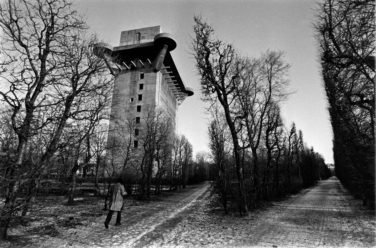Solitary figure beneath towering brutalist concrete architecture framed by bare winter trees in black and white.