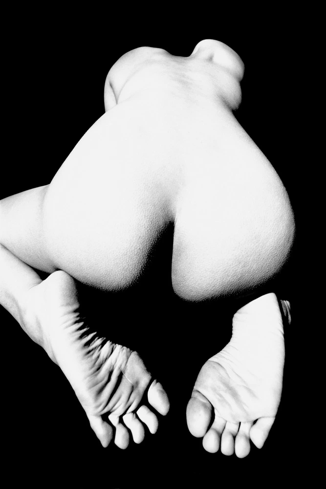 Abstract black and white nude photograph showing a crouched female figure transformed into sculptural form through dramatic high-contrast lighting against a black background.