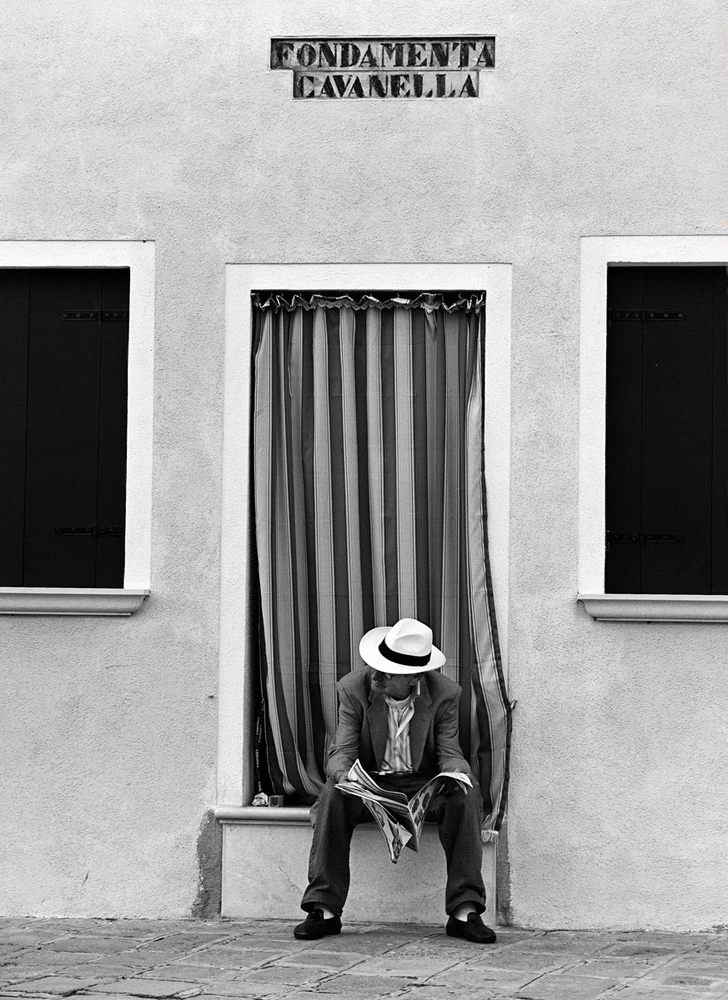 Black and white street photograph in Venice showing a man in a white hat reading a newspaper beneath the Fondamenta Cavanella sign, framed by striped curtains and minimalist architecture.