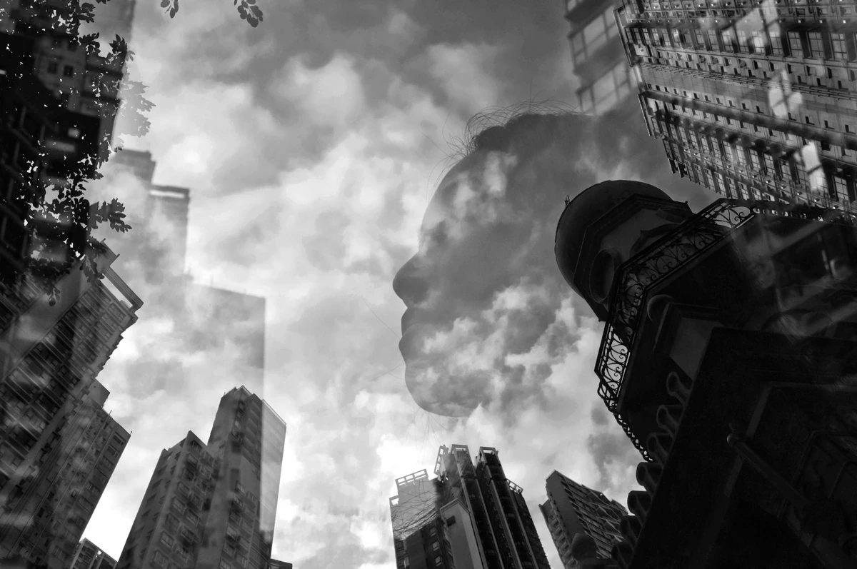Black and white multiple exposure portrait in Hong Kong featuring a woman's profile layered with dramatic clouds and towering city buildings, creating a dreamlike urban composition.