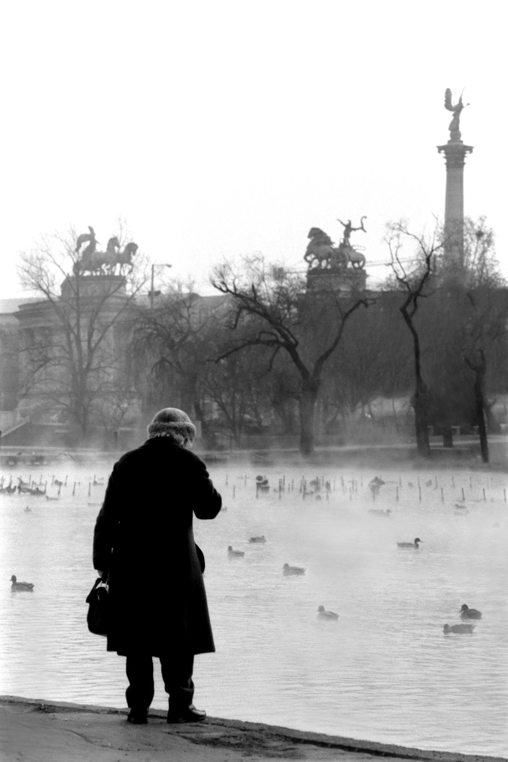 Elderly woman beside a pond in Budapest's City Park, looking towards Heroes' Square in black and white.